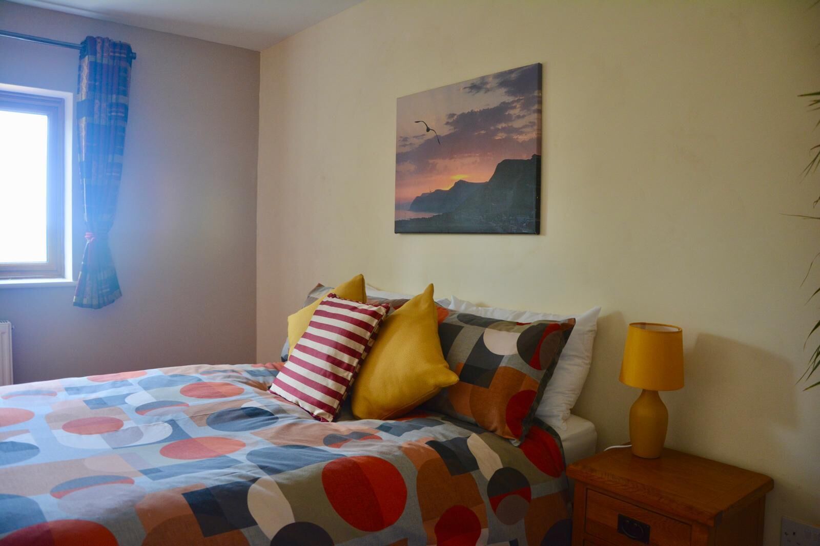 Une chambre à coucher avec un lit, des oreillers, une table de chevet, une lampe, une fenêtre avec des rideaux et un tableau au mur.