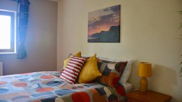 Une chambre à coucher avec un lit, des oreillers, une table de chevet, une lampe, une fenêtre avec des rideaux et un tableau au mur.
