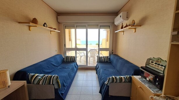 Una habitación con dos camas azules, una mesita con una radio y vista al mar desde la puerta.
