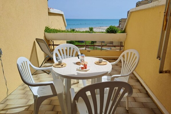 Un petit espace extérieur aménagé pour prendre les repas, avec une table et des chaises, une vue sur la plage et un mur jaune.