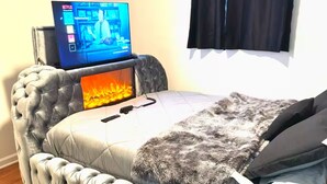 Un dormitori amb un sofà de reposacapses amb borlonas, una televisió de pantalla plana, una llar de foc i una finestra gran amb cortines.