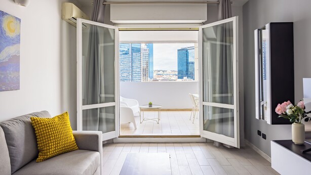 Un salón moderno con un sofá gris, un cojín amarillo y un ventanal de vidrio que da a un balcón exterior con vistas a la ciudad.