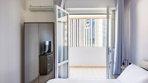 Una habitación de hotel moderna con una cama, un armario, un escritorio con lámpara y vistas a la ciudad a través de un ventanal grande.