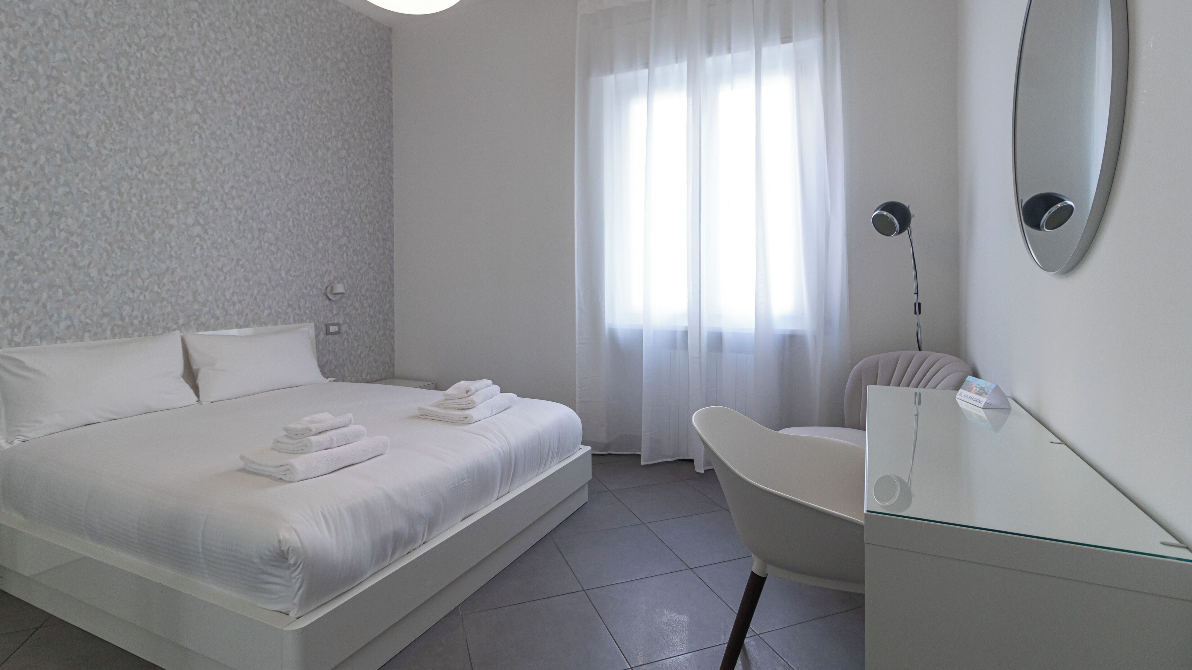 Une chambre d’hôtel moderne avec un lit bien fait, une chaise, un bureau et une grande fenêtre ornée de rideaux légers.