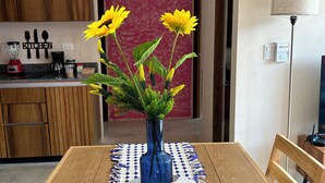 Un comedor con una mesa de madera, un florero azul con flores amarillas y una cocina al fondo.