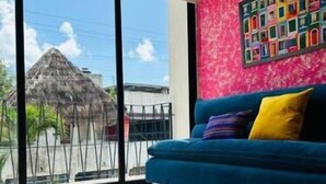 Una habitación con un sofá azul, un tapete estampado y un ventanal con vistas a un edificio y una construcción con techo de paja.
