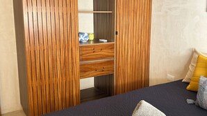 Un dormitorio con una cama azul oscuro, un armario de madera con aire acondicionado integrado y un estante pequeño con objetos decorativos.
