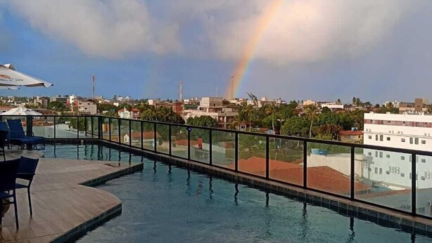 A swimming pool with a glass railing, a view of a cityscape, and a rainbow in the sky. - Flat no centro de Porto de Galinhas  (Ipojuca)