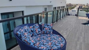 A round, woven outdoor seating area with a floral cushion pattern, located on a rooftop terrace with a glass railing and a view of the cityscape. - Flat no centro de Porto de Galinhas  (Ipojuca)