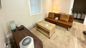 Una sala de estar moderna con una mesa de centro de madera, un sofá marrón con cojines, una ventana con persianas y una lámpara pequeña.