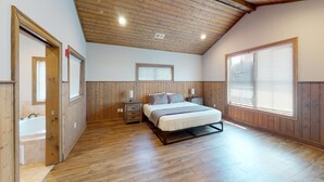 Een slaapkamer met een houten plafond, een groot raam met zonwering, een bed met matras en kussens, een nachtkastje met een lamp en een houten deur.