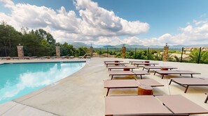 A poolside area with lounge chairs and tables, a clear pool, and a mountainous backdrop.