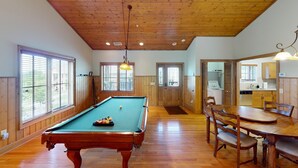 Una mesa de billar en una habitación con pisos y paredes de madera, una mesa de comedor con sillas y una zona de cocina visible a través de una puerta abierta.