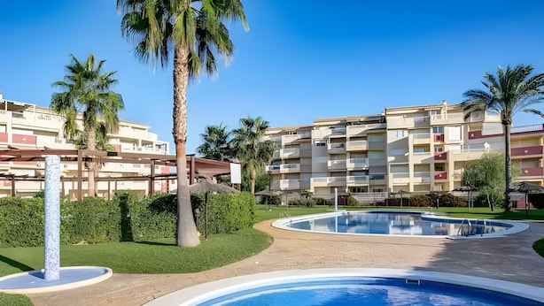 A swimming pool area with palm trees and a multi-story building in the background. - Playasol in Denia With 3 Bedrooms and 2 Bathrooms (Denia)