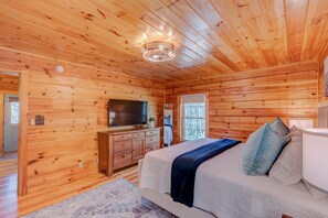 Ein Schlafzimmer einer Holzhütte mit einem Bett, einem Fernseher auf einer Holzkommode und einem Fenster mit Blick auf Bäume.