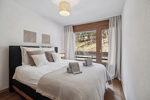 Uma cama bem arrumada com cabeceira, uma janela grande com cortinas e duas obras de arte fixadas na parede.