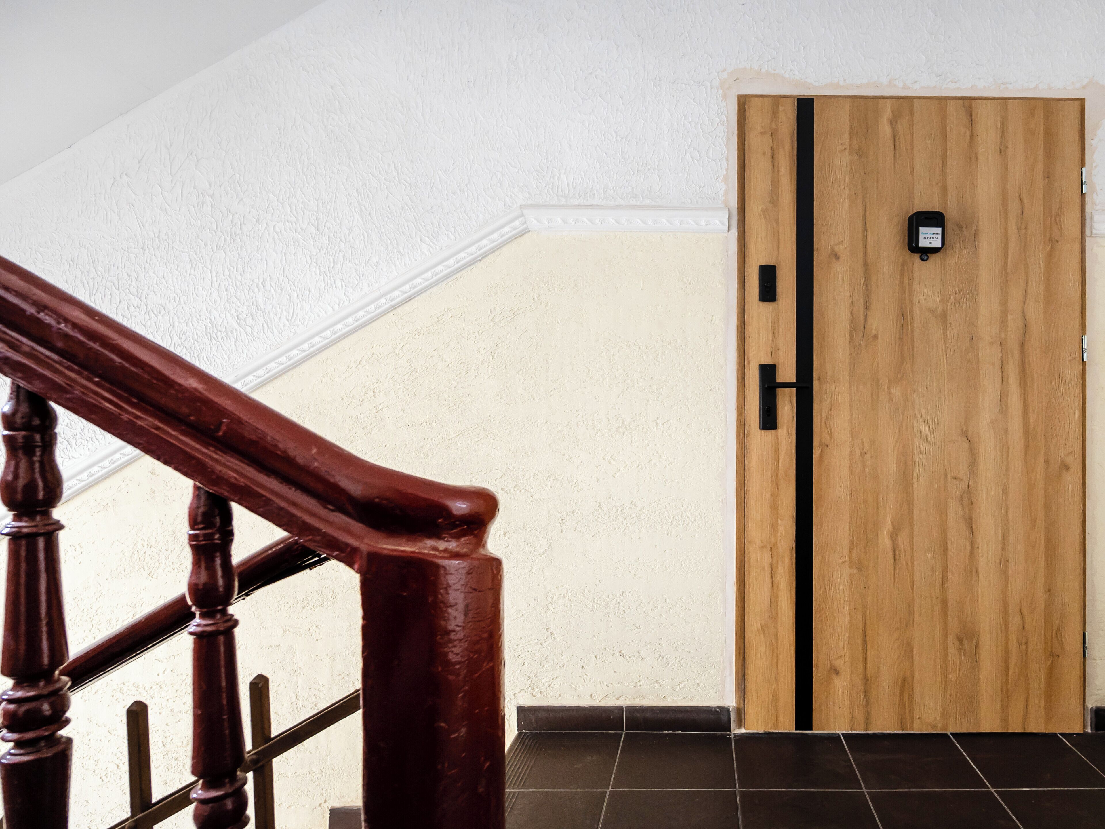 Une porte en bois dotée d’un clavier pour l’ouverture, un escalier en bois avec une rampe et un sol carrelé.