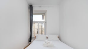 A narrow hotel room with a single bed, white linens, and a view of buildings through a window. - Kilińskiego Standard Apartment (Łódź)