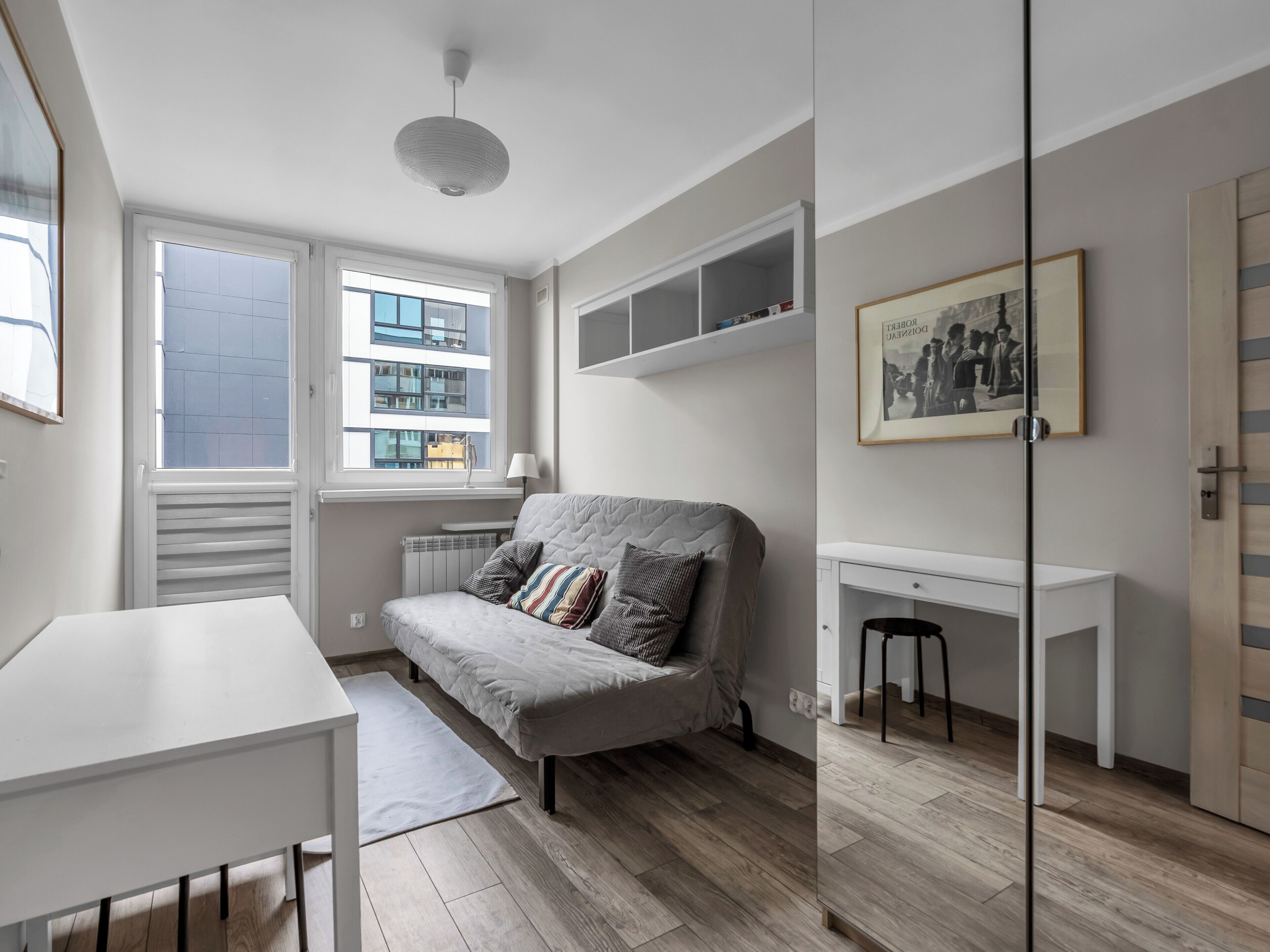 Une pièce moderne et compacte, avec un canapé gris, un bureau blanc et un radiateur. Une fenêtre laisse entrer la lumière naturelle, et un tableau encadré est accroché au mur.