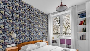 Ein ordentlich eingerichtetes Schlafzimmer mit gemustertem Tapeten, einem hölzernen Bett, weißer Bettwäsche, einem Nachttisch, einem Fenster mit Blick auf Bäume und einem Bücherregal.