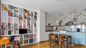 Eine moderne Küche mit Holzparkett im Fischgrätenmuster, einem Essbereich mit rundem Tisch und Stühlen sowie einem weißen Bücherregal voller Bücher und Dekoartikeln.