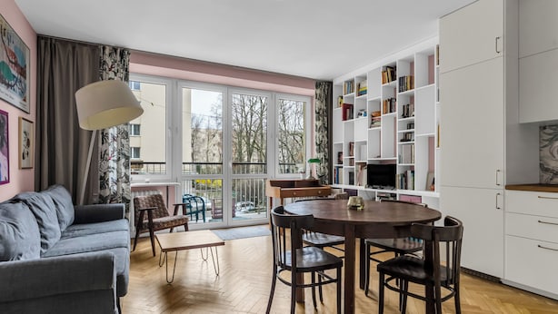 Ein modernes Wohnzimmer mit grauem Sofa, einem Holzesstisch, Stühlen, einem Bücherregal und einem großen Fenster mit Blick auf Bäume.