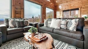 A cozy living room with a large wooden ceiling, two sofas with patterned cushions, a wooden coffee table with a tray of snacks, and large windows offering a view of the outdoors. - Top of the World: Summer deals-Theater-hot tub-arcade games-million dollar views (Sevierville)