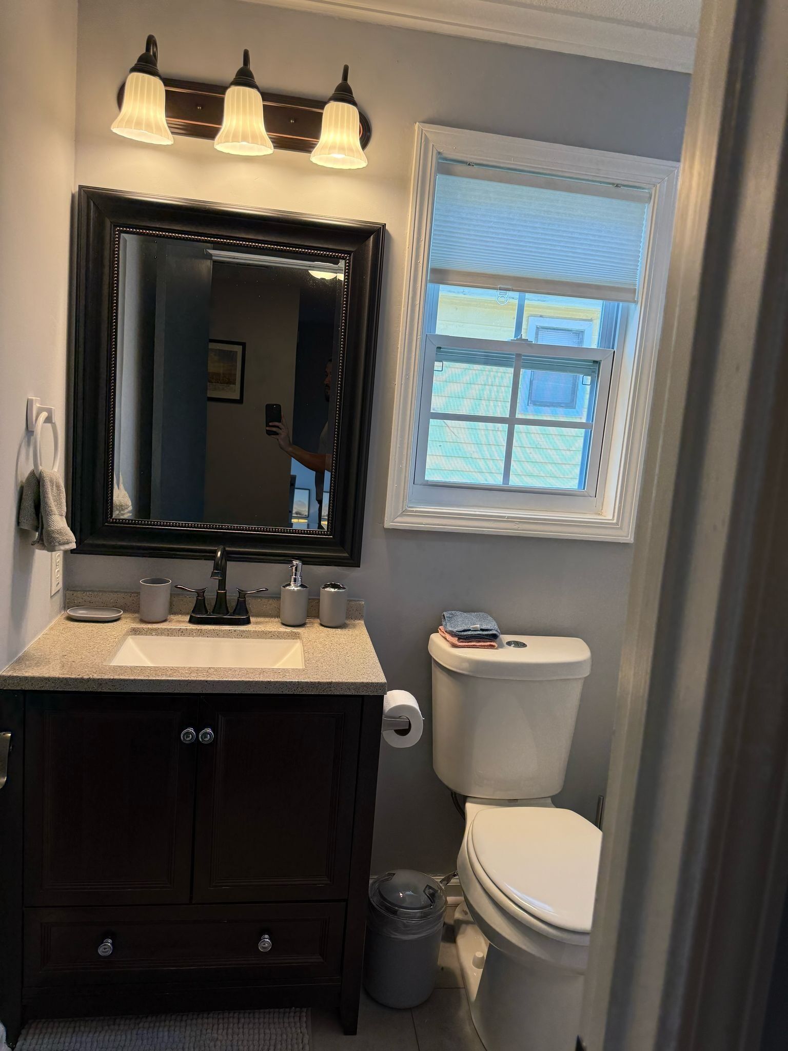A bathroom with a dark wood vanity, a large mirror, a sink, and a toilet. There is a window above the toilet and a trash can next to it.