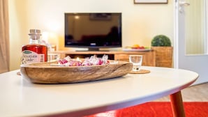 Un cuenco de madera con aperitivos y una botella de bebida roja sobre una mesa blanca en una habitación con televisión y puerta.
