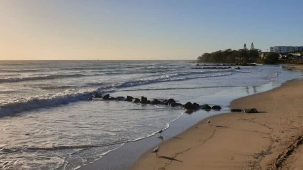 A beach with waves, a sandy shore, and buildings in the background. - Beachside on Burkitt - duex (Bargara)