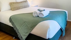 A neatly made bed with a green throw blanket, white pillows, and rolled towels. There is a framed picture of a beach scene on the wall. - Beachside on Burkitt - duex (Bargara)