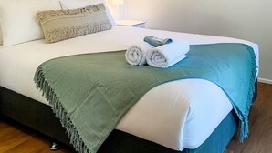 Een net opgemaakt bed met een groene deken, witte kussens en een ingelijst foto van een strand.
