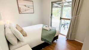Une chambre à coucher comprenant un lit, une table de chevet, un ventilateur de plafond, une fenêtre avec des rideaux et une porte donnant sur un espace extérieur.