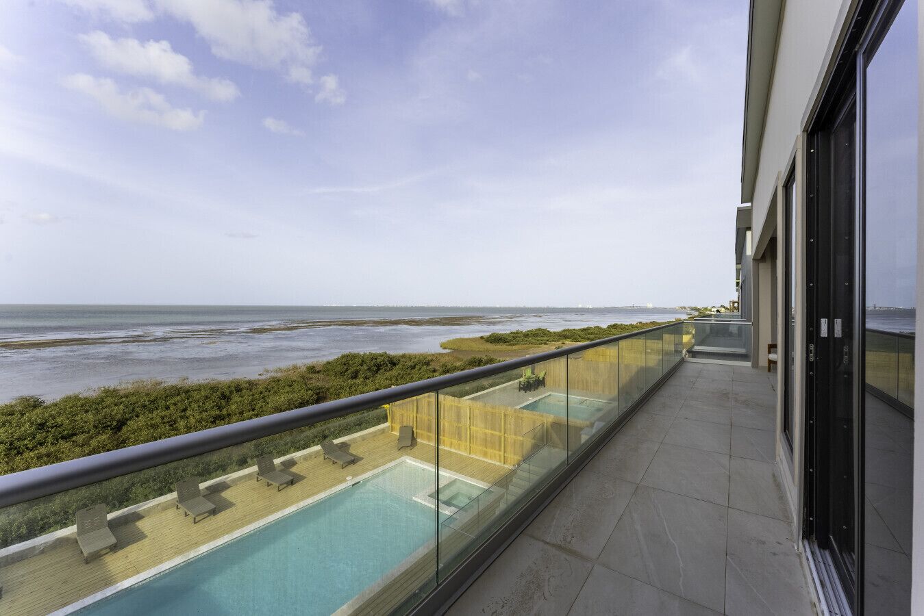 A balcony with a glass barrier overlooking a swimming pool and the sea.