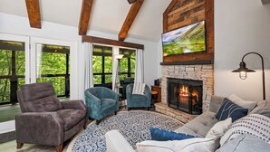 A cozy living room with a stone fireplace, wooden beams, and large windows overlooking greenery.