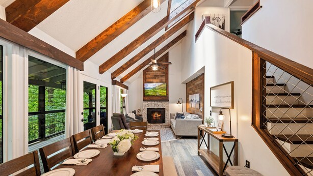 A dining area with a long wooden table set for six, a fireplace, and a staircase with a wooden railing.