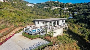 Une maison moderne dotée d’un grand patio, d’une piscine et offrant une vue sur une colline où se trouvent d’autres maisons.