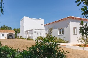 A white building with a red-tiled roof, a covered car parking area, and surrounding greenery.