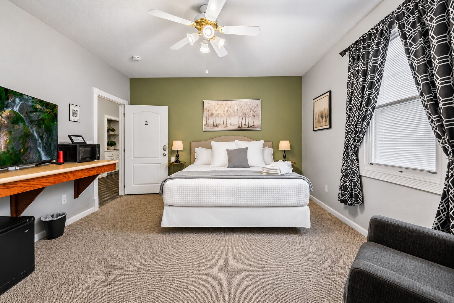 A bedroom with a bed, a desk, a television, a ceiling fan, and artwork on the walls.