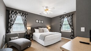 Une chambre à coucher comprenant un lit, deux tables de chevet, une chaise, un bureau, un ventilateur de plafond et deux fenêtres avec des rideaux.