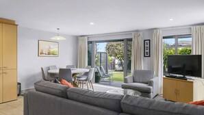 A modern living room with a grey sofa, a dining table with chairs, a TV, and a sliding glass door leading to a garden. - 3 BR Holiday home - Steps to the lake and short, scenic stroll to town (Taupō)