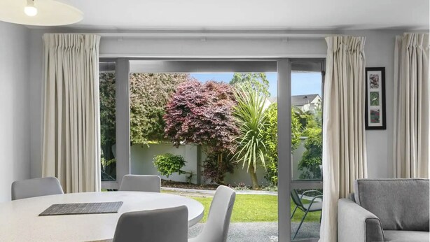 A modern living room with a round table and chairs, a grey sofa, and a sliding glass door leading to a garden with trees and a lawn. - 3 BR Holiday home - Steps to the lake and short, scenic stroll to town (Taupō)