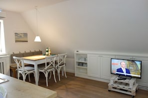 Uma sala de jantar com mesa de madeira e cadeiras brancas, uma televisão em suporte branco e um armário branco com prateleiras.
