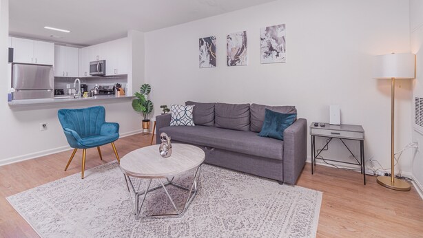Una sala de estar moderna con un sofá gris, un sillón azul y una mesa de centro redonda. Se ve una cocina al fondo con armarios blancos y electrodomésticos de acero inoxidable.
