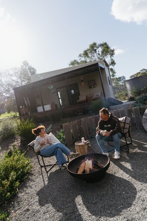 Zwei Personen sitzen an einer Feuerstelle in einem Garten, im Hintergrund ein modernes Haus.