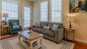 A living room with a sofa, a coffee table, and a lamp. There are two windows with blinds, a painting on the wall, and a door.