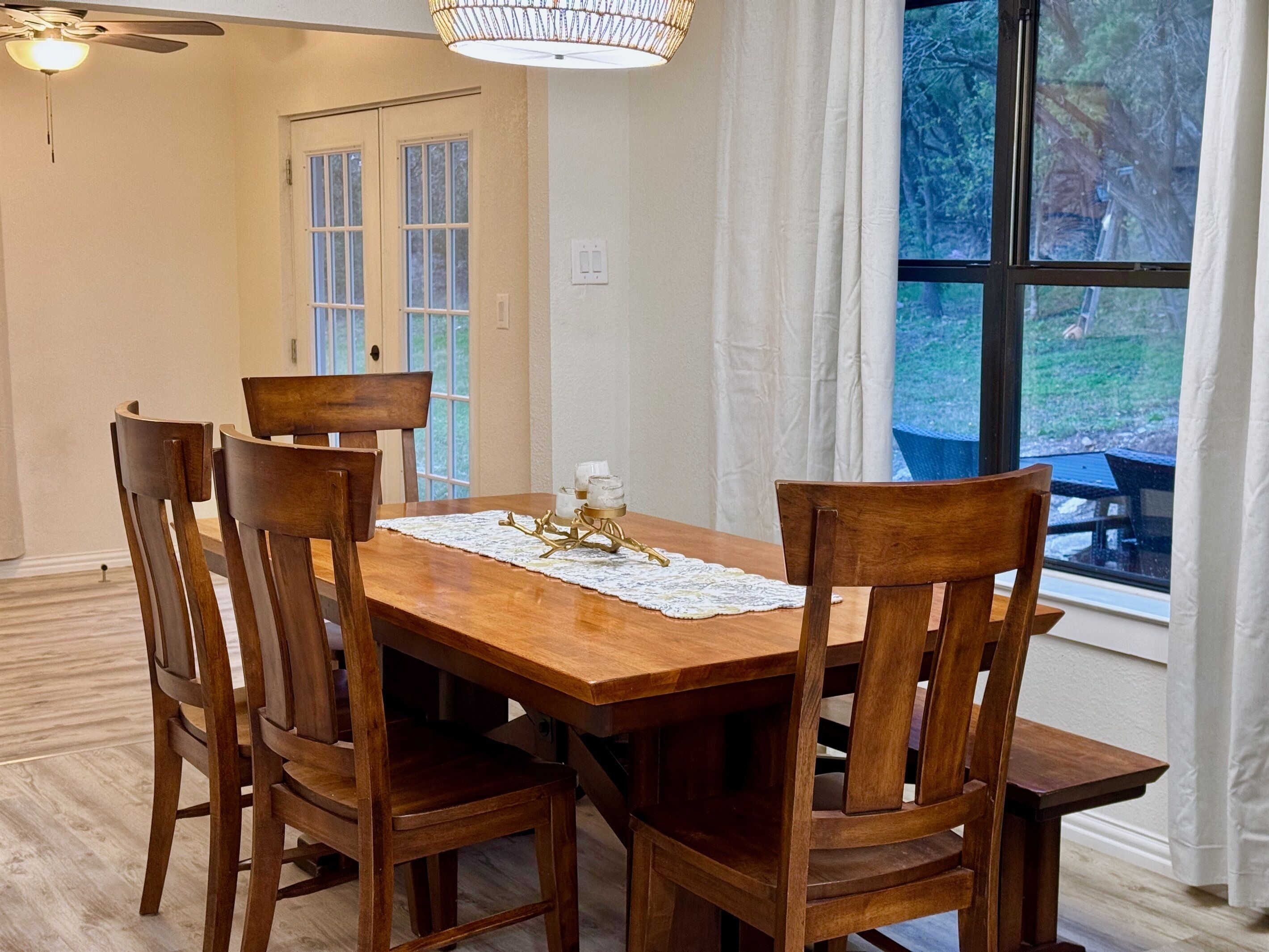A dining area with a wooden table and chairs, a lace table runner, and a view of the outdoors through a large window.