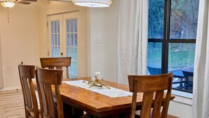 A dining area with a wooden table and chairs, a lace table runner, and a view of the outdoors through a large window. - Hilltop Harbor House (Granbury)