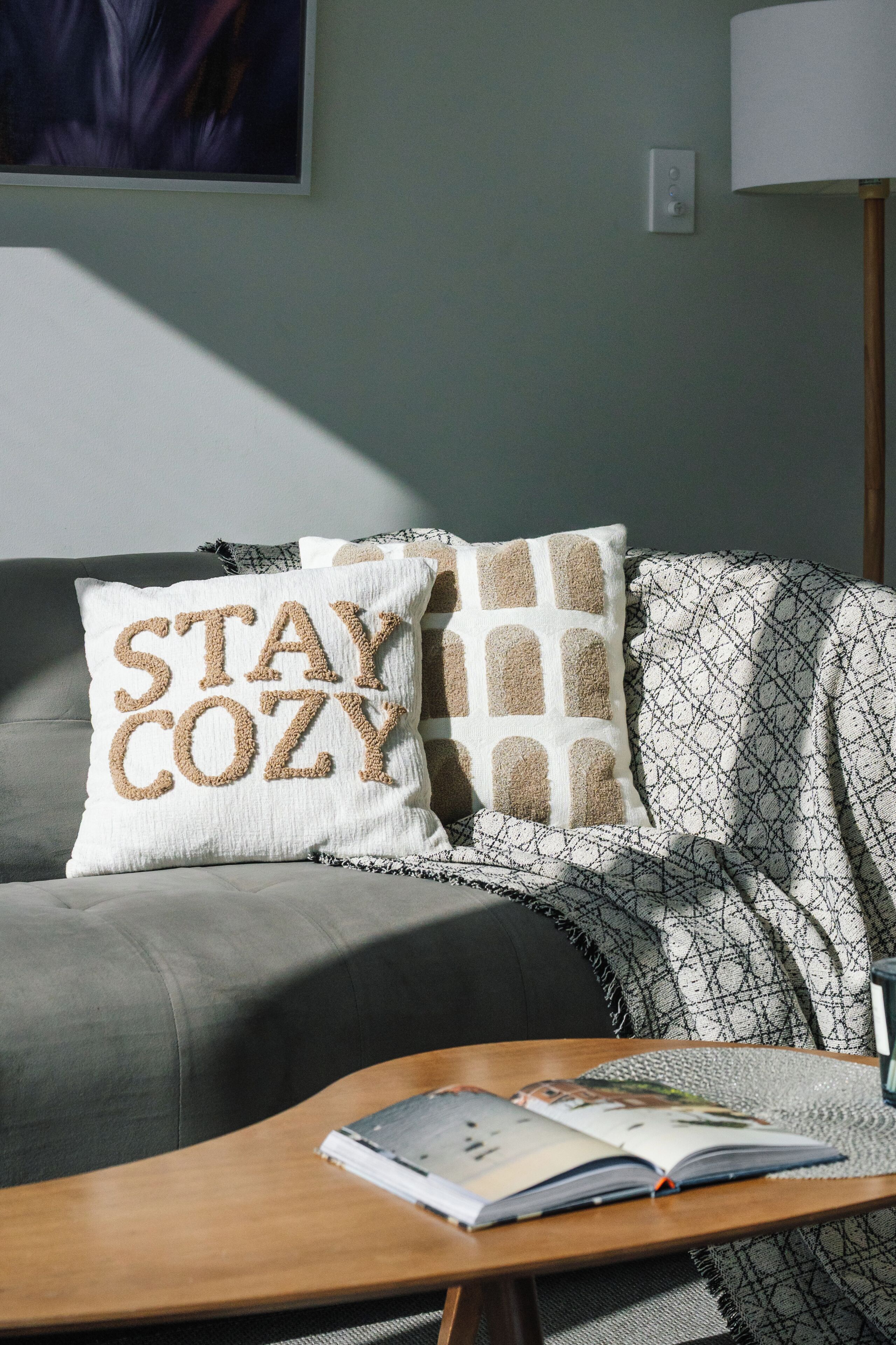A living room with a grey sofa, a 'STAY COZY' pillow, a patterned throw, and a wooden coffee table with an open magazine.