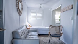 Ein Zimmer mit einer weißen Ledersofa, einem Korbstuhl, einem Bett mit weißer Bettwäsche, einem Deckenventilator und einem Schild mit der Aufschrift 'BEACH HOUSE'.